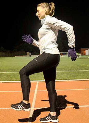 Rapariga em posição estática, realiza técnica de corrida, na pista de atletismo, à noite.