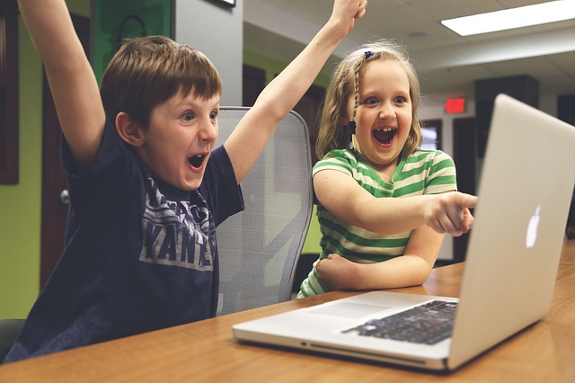 Crianças, festejam, junto a secretária, olhando para o ecrã do computador portátil.